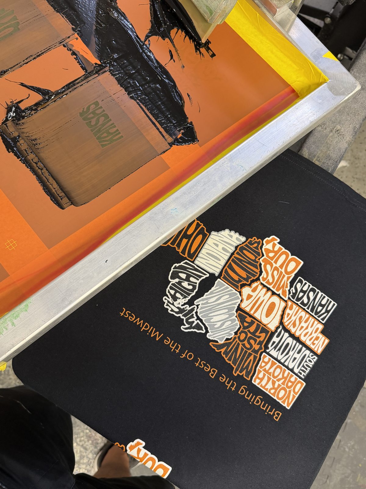 An inked screen and squeegee on the press above a black shirt with a multicolor Midwest graphic.